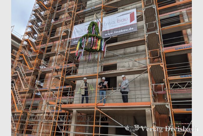 Kein Richtfest ohne Richtbaum: Bunt geschmückt hing dieser an der Vorderseite des ersten Bauabschnitts des neuen Wohnquartiers An der Ölmühle. Hawobau-Geschäftsführer Holger Kazzer (links) und Bürgermeister Klaus Schindling (rechts) lauschen dem Richtspruch, den in Vertretung eines Zimmermanns Patrick Vogel von der f.m.a. bau GmbH verlas.